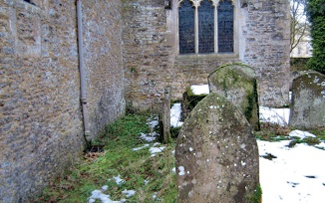 A shallow depression which is a few feet wide separates a church wall from a partially snow-covered graveyard