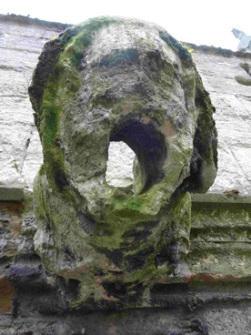 Eroded gargoyle with green biological growth; the form is still just recognisable as a human head