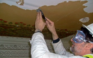 A conservator hand-strips white emulsion from a ceiling to reveal a historic paint scheme with an elaborate flower and trellis-work border