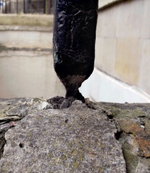 Close-up of cracked and spalled stone plinth around junction with heavily rusted iron paling