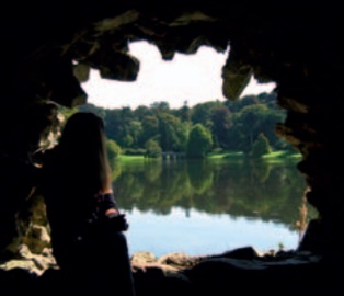 Woman looking out from grotto at view over lake to stone bridge and parkland on further shore