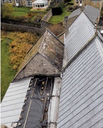 Tarpaulins cover a flat area of church roof tucked behind a pitched, slated roof; houses are visible in the background