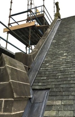 Wide lead channel at junction of slate roof and stone gable parapet and lead roll protecting slate edges