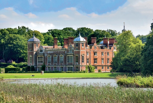 Red-brick country house with square corner turrets; lake and lawns in foreground