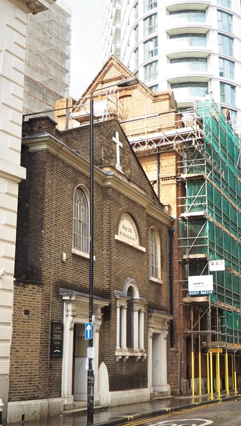 The Georgian brick facade of St George's Lutheran church in Aldgate, bracketed by taller buildings and with skyscrapers looming in the background
