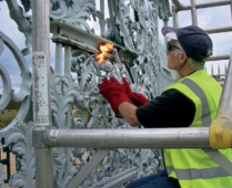 A heritage ironwork student in a high-visibility vest flame cleans an ornate ironwork screen