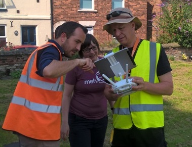A man in a high-vis jacket uses a remote control device to direct a drone while two other people look at the screen relaying the camera footage